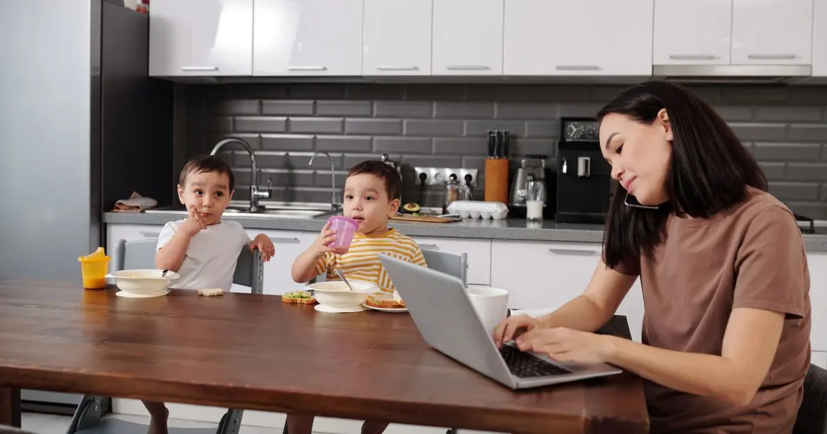 A mother on a phone call typing on her laptop at the kitchen table while her two young sons eat breakfast.