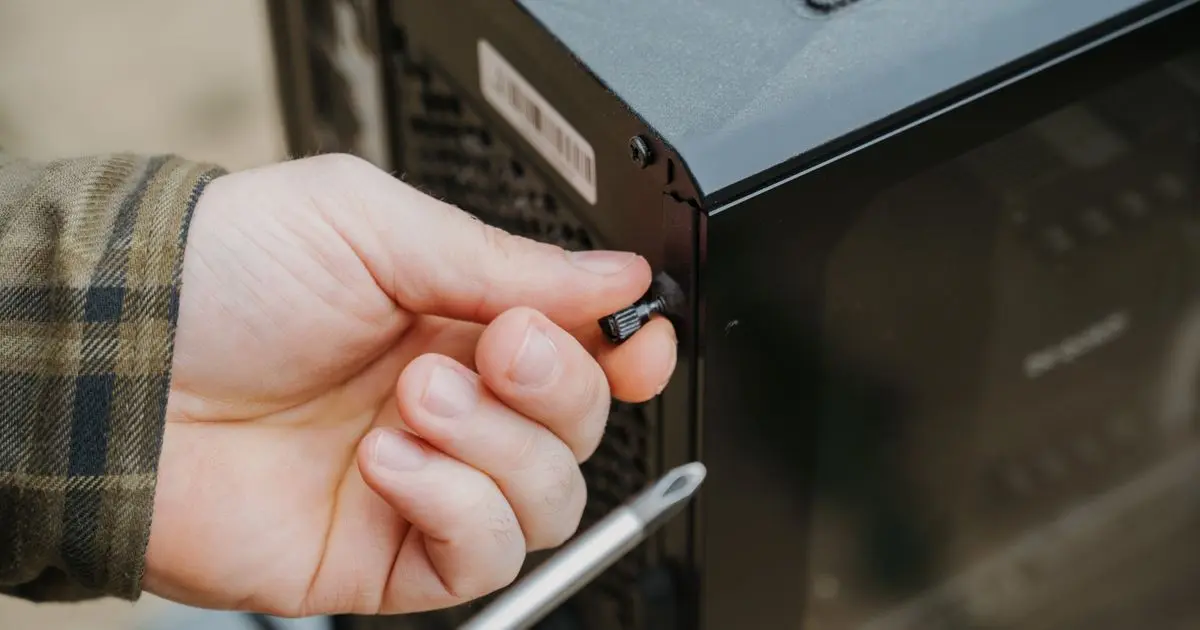 A hand loosening the thumbscrew on a desktop computer case, a screwdriver beside it.
