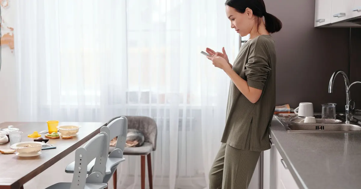 A woman in her kitchen in the morning, checking her phone by the counter.