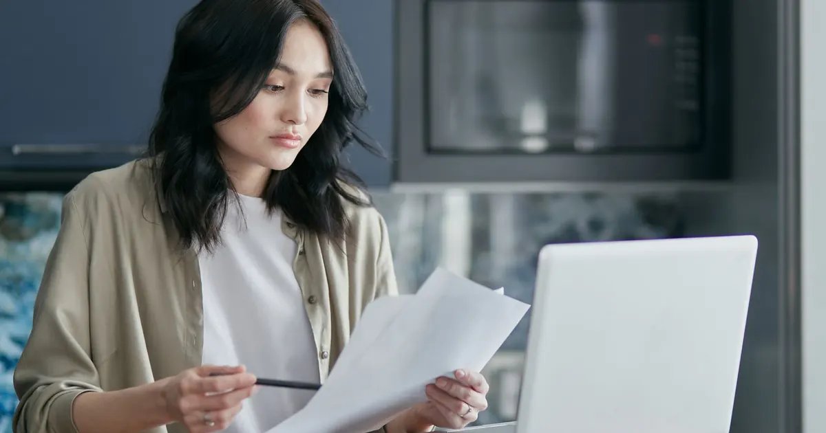 A woman at her kitchen counter reading papers with a pen in hand, her laptop open beside them.