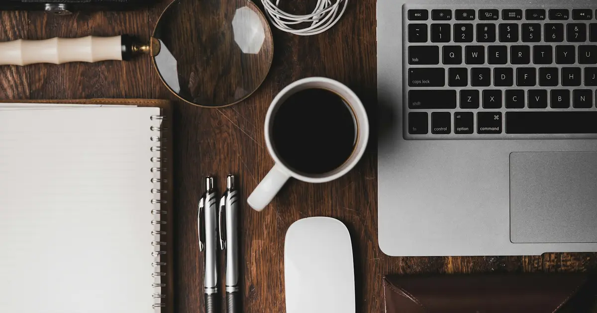 An overhead view of a tidy desk with a notebook, pens, a laptop, and a cup of coffee.
