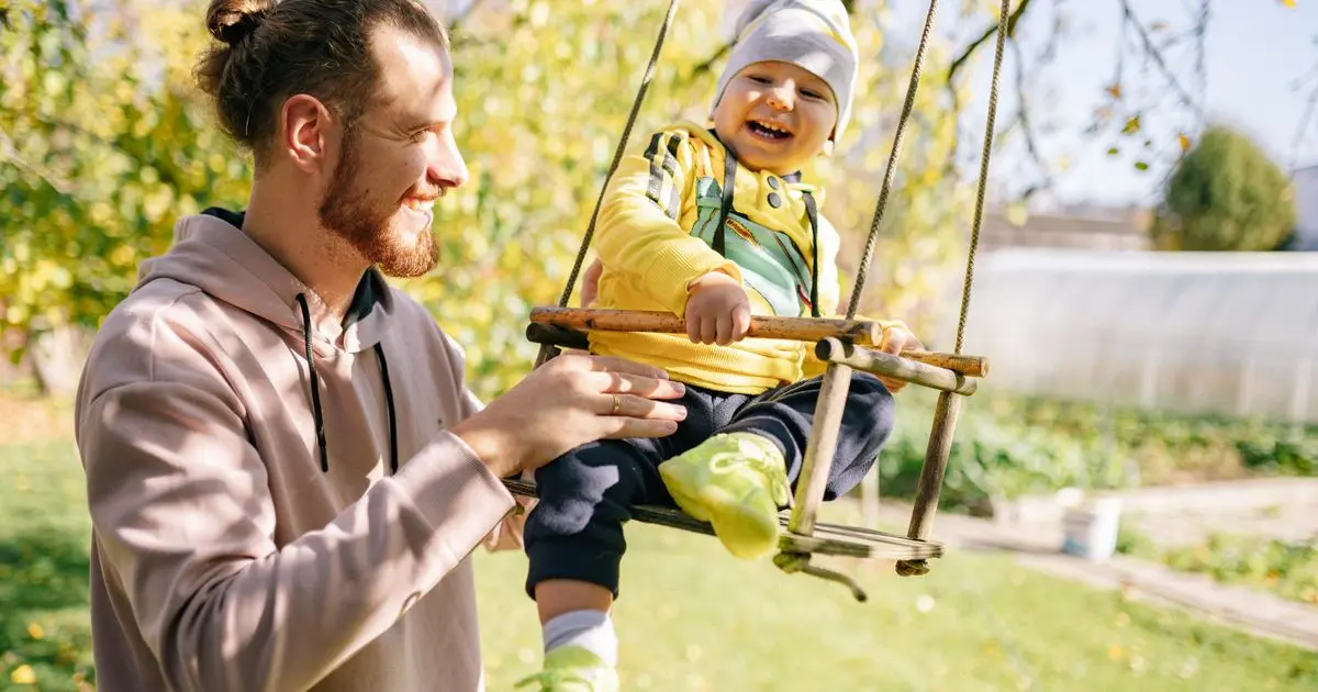 A smiling father pushing his laughing toddler on a wooden swing in the backyard.