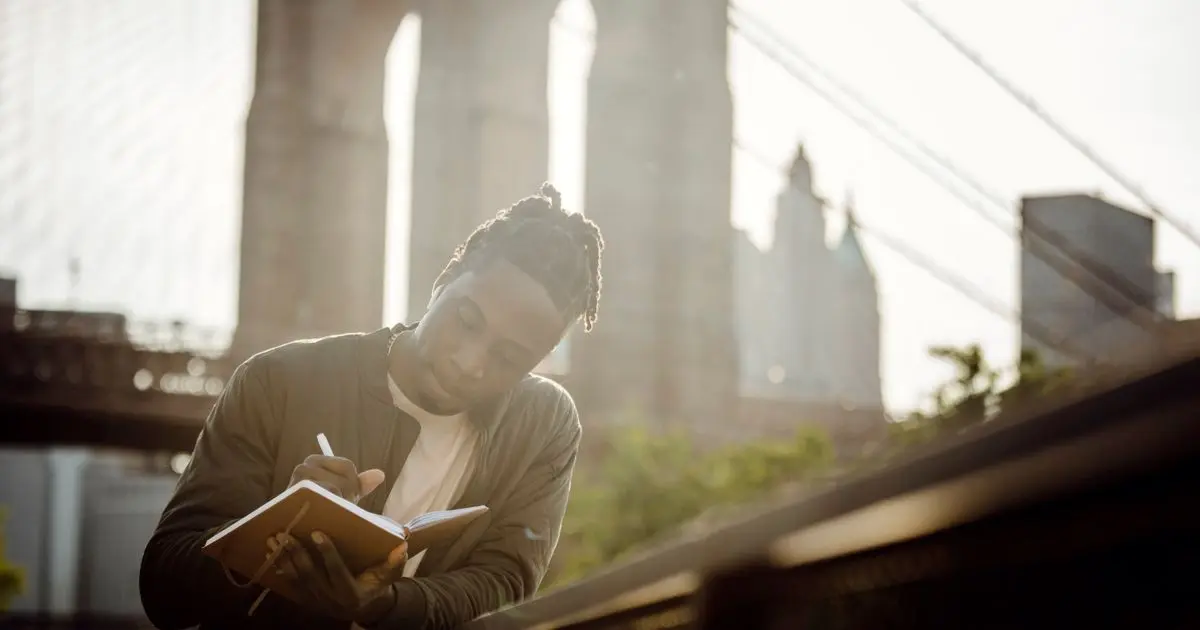A man writing an idea in his notebook outdoors, with a bridge behind him.