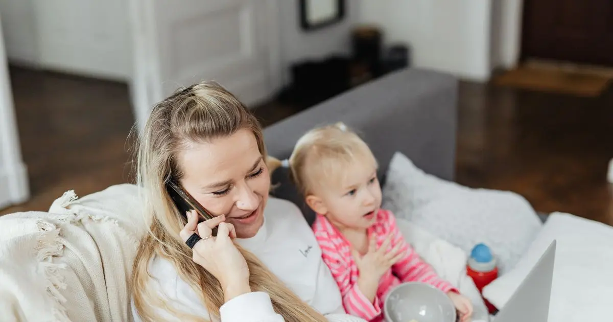 A mother on a phone call on the couch while her toddler looks up at her.