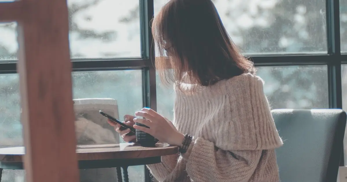 A woman sitting by a rainy window with her coffee, glancing at her phone.