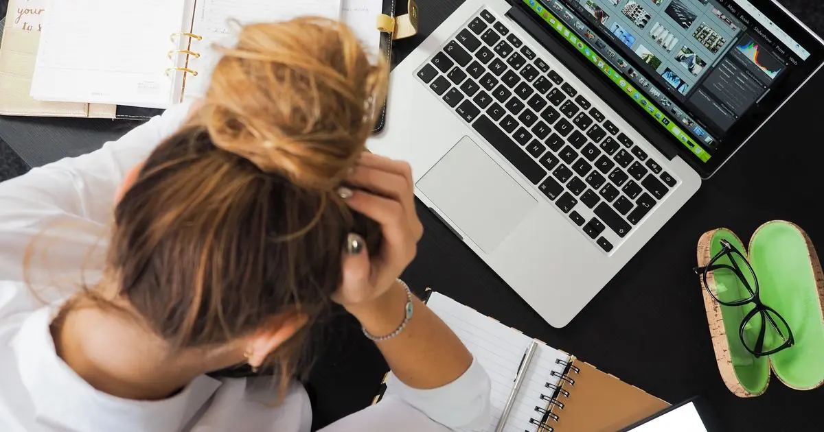 An overwhelmed woman at a desk with her hands in her hair, surrounded by a laptop, a planner and a phone.