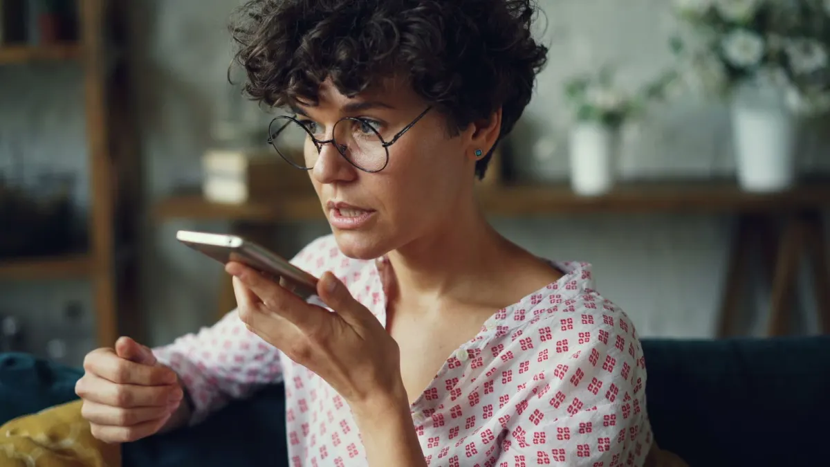 A mom at home in a casual blouse, relaxed, speaking a voice memo into her phone to plan her day.