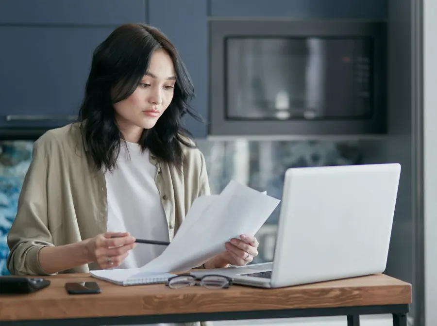 A woman at her kitchen counter reading school papers with a pen in hand, her laptop open beside them.
